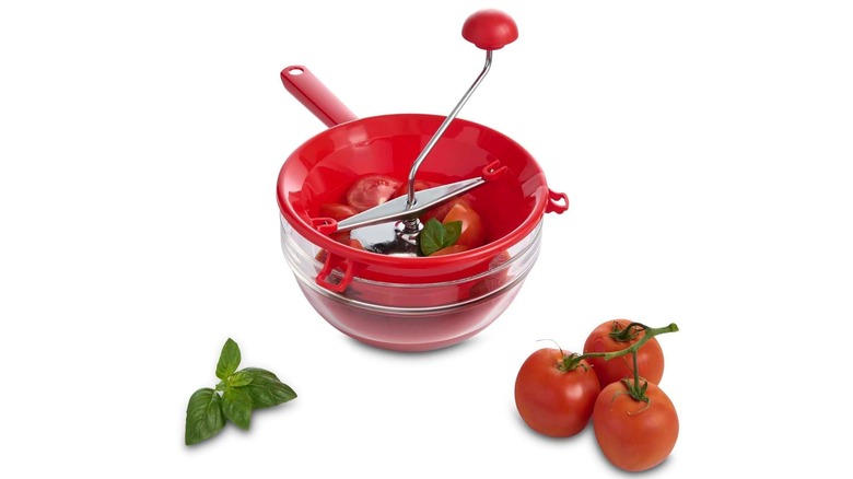 A red plastic and steel food mill with tomatoes and basil inside
