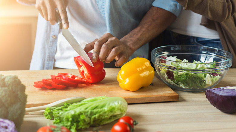 A person chopping a red bell pepper on a wooden cutting board among other salad vegetables.