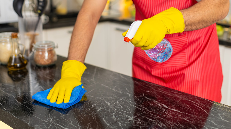 A person wearing a red apron and yellow gloves spraying cleaning solution on a kitchen counter and wiping it down