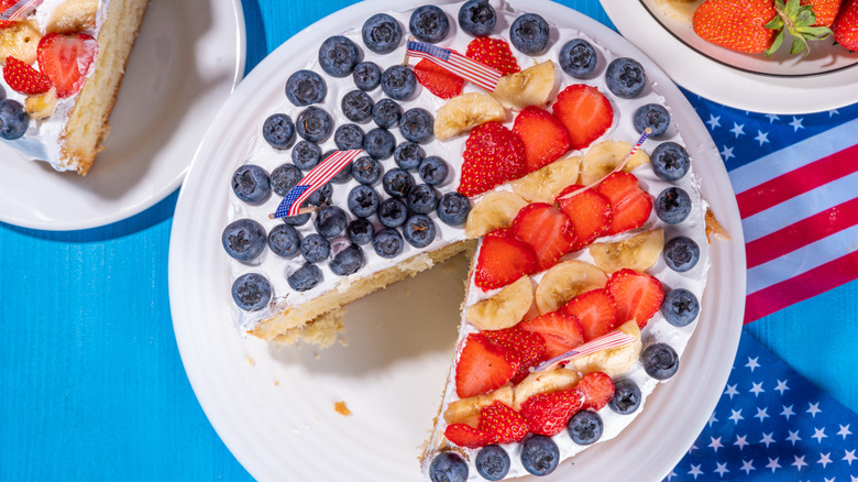 Yellow cake with white icing and American flag design made out of blueberries, strawberries, and bananas