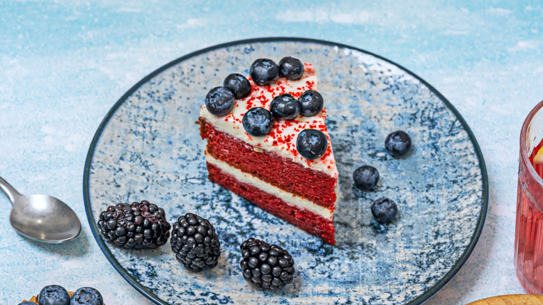 A slice of red velvet cake with white icing and blueberries on top, plus blackberry garnish