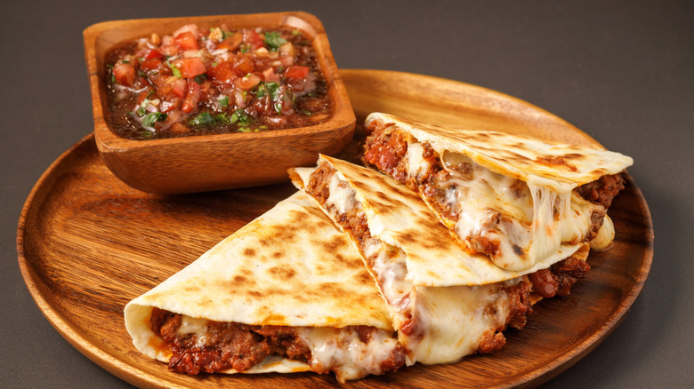 A meat and cheese quesadilla on a wooden plate with a side wooden bowl of salsa