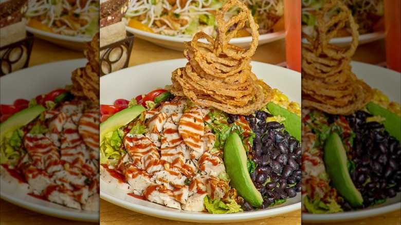 A salad with lettuce, tomatoes, black beans, corn, avocado, chicken, and a tower of onion rings