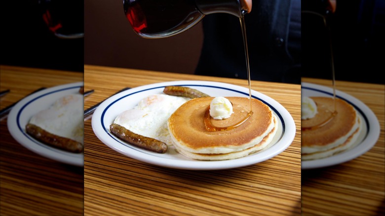 Syrup pouring over two pancakes with butter, two sausage links, and two eggs
