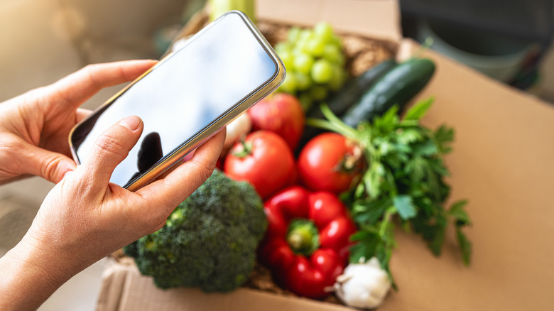 A person using their phone in front of a box of fresh vegetables