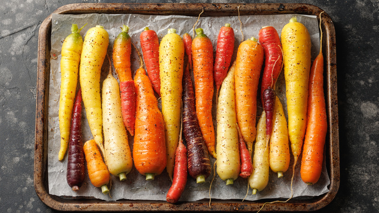 A row of differently colored carrots on a baking sheet and covered with seasoning