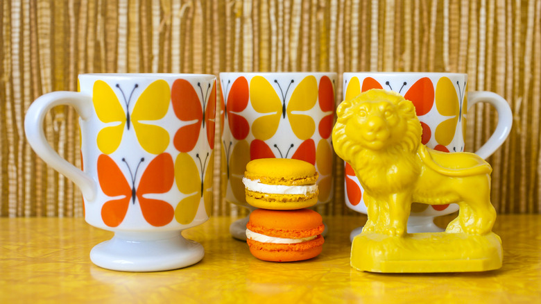Three white mugs with orange and yellow butterflies alongside lion decoration and macarons