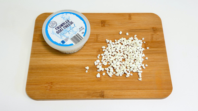 Pieces of cheese on a cutting board next to a container of Emporium Selection Original Crumbled Goat Cheese