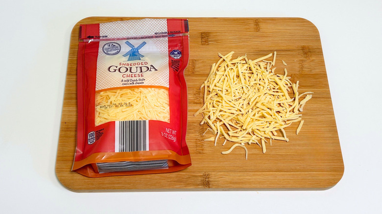 Pieces of cheese on a cutting board next to a bag of Emporium Selection Shredded Gouda Cheese