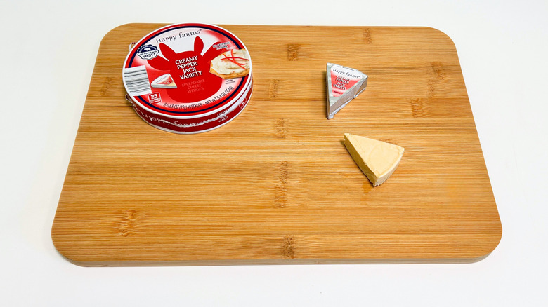 Pieces of cheese on a cutting board next to a container of Happy Farms Creamy Pepper Jack Variety Spreadable Cheese Wedges