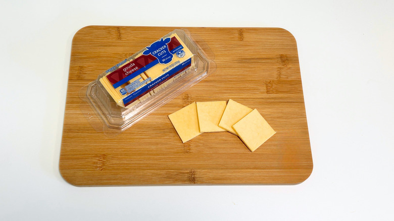 Pieces of cheese on a cutting board next to a container of Happy Farms Gouda Cheese Cracker Cuts