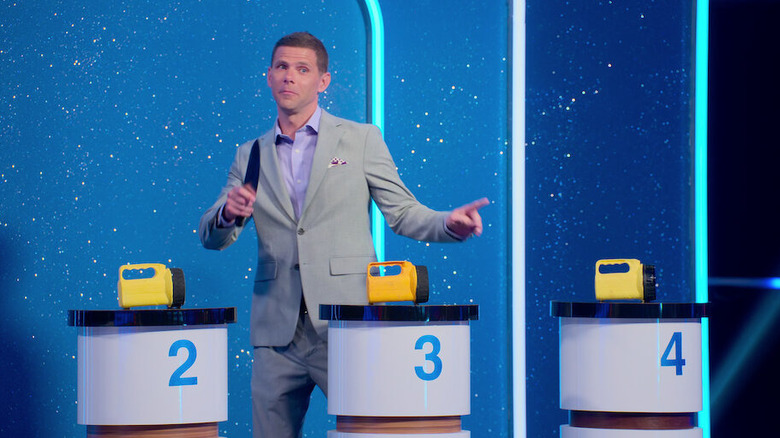 "Is It Cake?" host Mikey Day standing in front of three podiums with flashlights with a knife in his hand