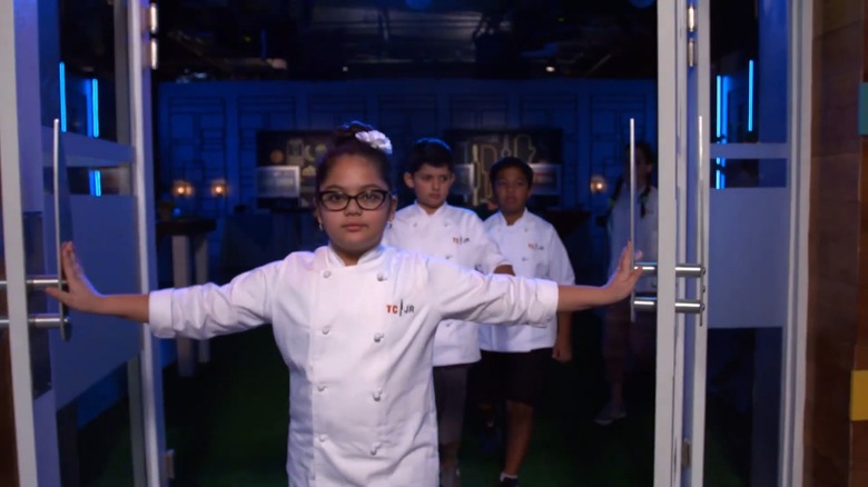 4 small children in white chef's jackets that say "TCJR" dramatically walking through a doorway