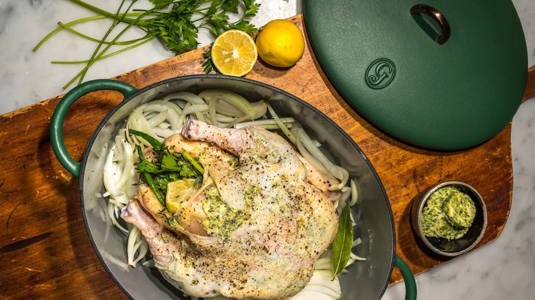 A green Dutch oven with a whole chicken inside on a wooden cutting board with lemons, herbs, and compound butter