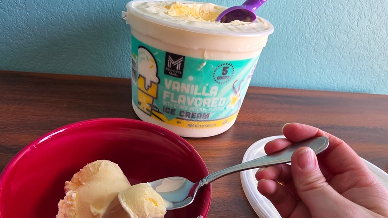 Member's Mark vanilla ice cream pail with ice cream scoop and hand dipping spoon into ice cream bowl