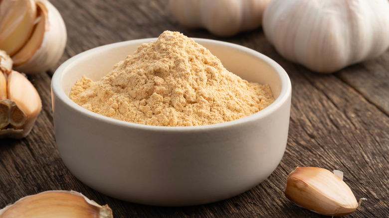 A white bowl with a mountain of garlic powder on a wooden table, surrounded by garlic cloves