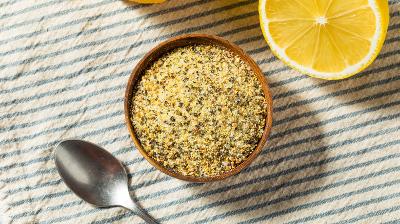 Lemon pepper seasoning in a small wooden bowl on a striped tablecloth with a lemon half and spoon next to it