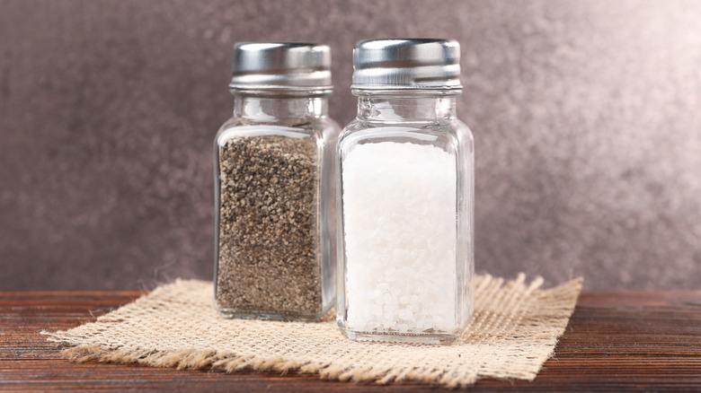 Two shakers, one filled with ground salt the other ground pepper, on a small woven coaster atop a wooden table