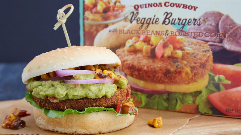 a veggie burger topped with lettuce, guacamole, red onion, and corn salsa in the foreground and a box of Trader Joe's Quinoa Cowboy Veggie Burgers with Black Beans & Roasted Corn in the background