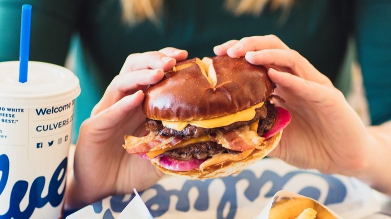Two hands holding a pub burger with a pretzel bun with a Culver's drink cup visible