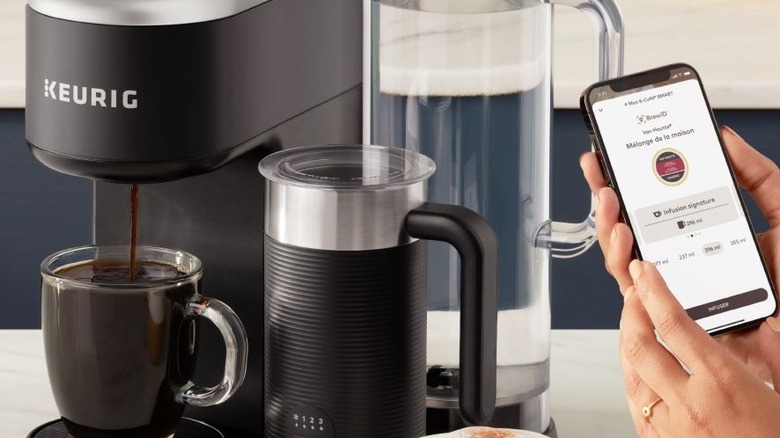 Keurig coffee machine pouring coffee into mug with hand holding phone with Keurig app open