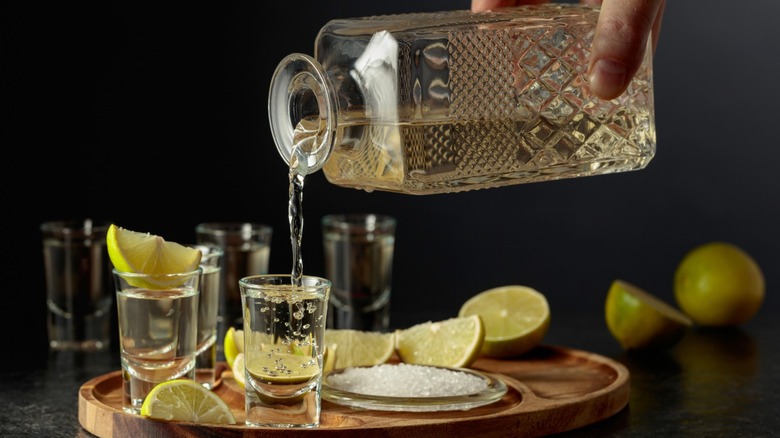 A person pouring tequila into a shot glass on a tray next to salt and limes.