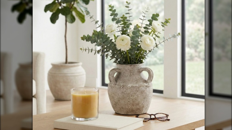 An off-white vase with white flowers next to reding glasses and a candle