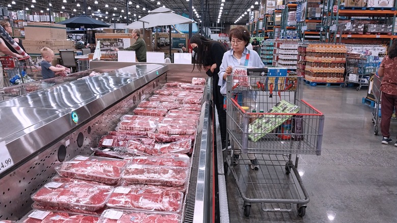 A woman pushing a shopping cart browses a meat refrigerator at Costco
