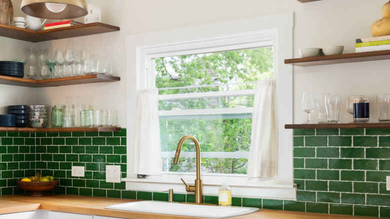 A bright window with opened café curtains in a kitchen with open shelves, wood countertops, and a green tile backsplash