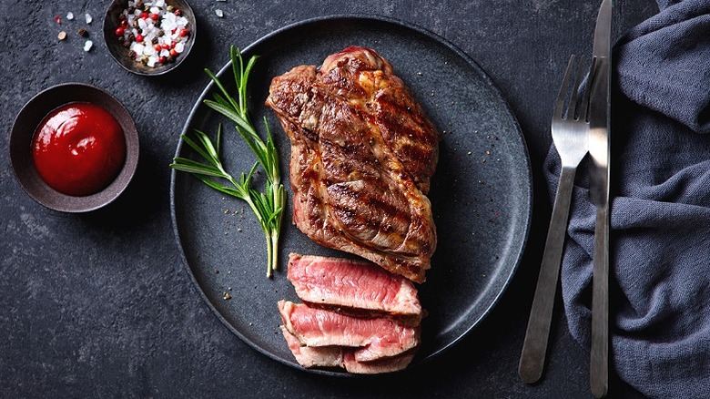 Beef Steak medium rare on a black plate, top view, with ketchup and seasonings ramekins and utensils to the side