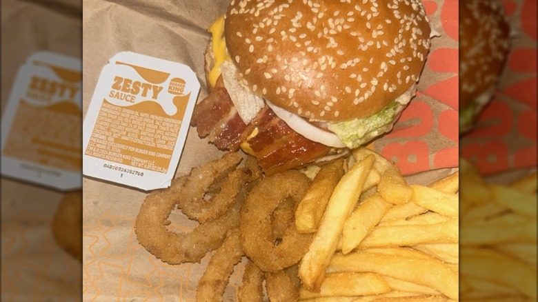 A Burger King bacon burger, french fries, and onion rings on paper with a sealed container of Zesty Sauce