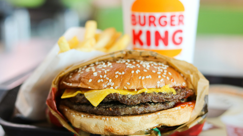 A close up of a Burger King cheeseburger in a wrapper with fries wand a branded soda cup in the background