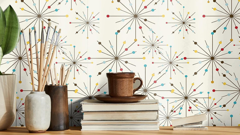 A desk with books, paintbrushes, and a mug on it in front of a wall covered in mid-century modern atomic starburst-style wallpaper