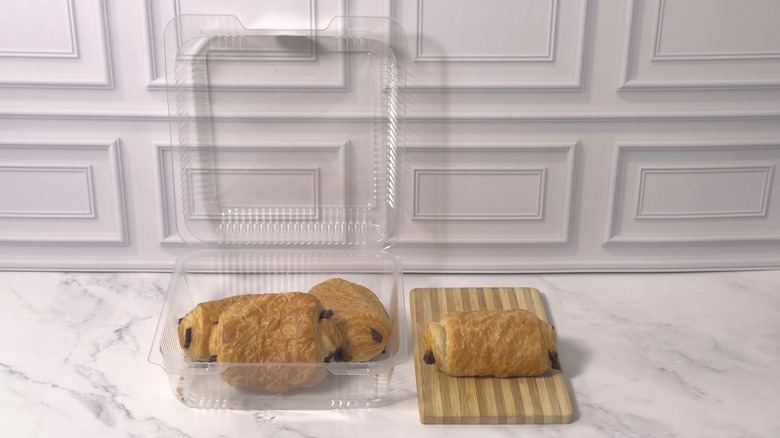 Clear plastic container of Whole Foods chocolate croissants and a whole croissant on a wooden cutting board