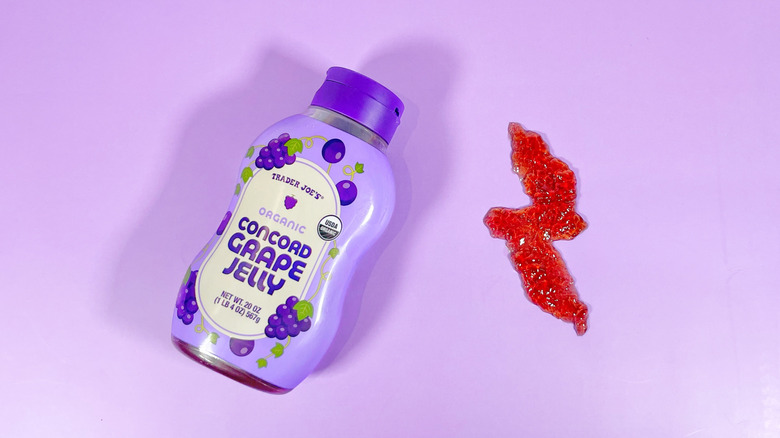 A plastic bottle of Trader Joe's grape jelly next to a zig zag of the jelly on a purple background