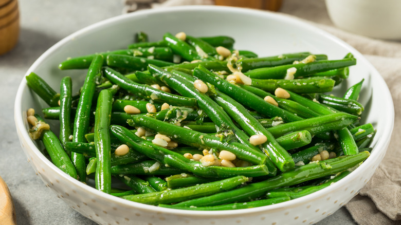 White bowl of cooked green beans garnished with pine nuts