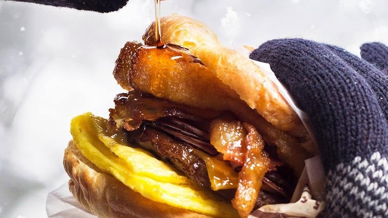 A hand pouring syrup onto breakfast sandwich with French toast sticks in it