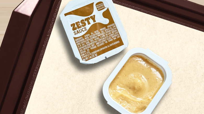 A sealed Burger King zesty sauce container next to an open container on a fast food tray
