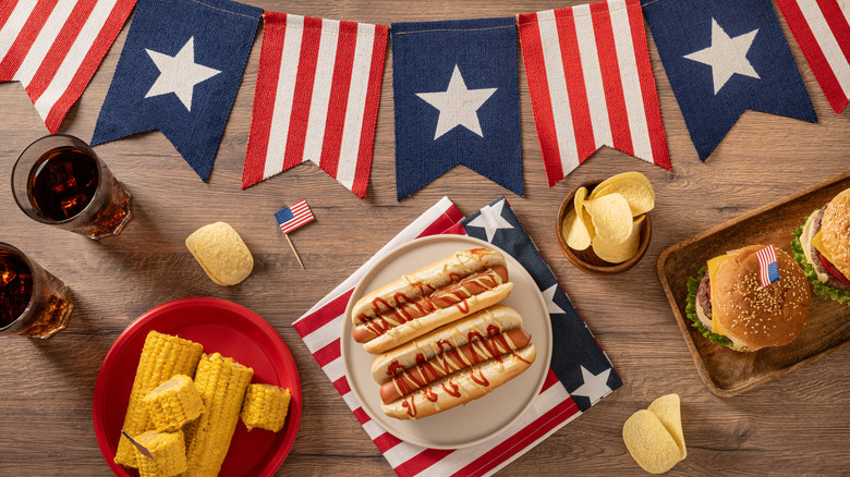 A food spread with an American flag banner, hot dogs, potato chips, corn on the cop, cheeseburgers, and soda