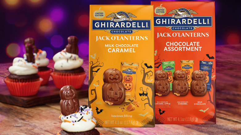 Two bags of Jack O'Lantern chocolates on a table with Halloween cupcakes, which are also decorated with the pumpkin shaped chocolates