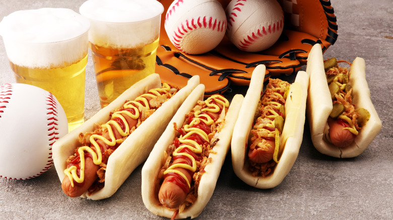 several hot dogs on surface along with baseballs, glove, and glasses of beer