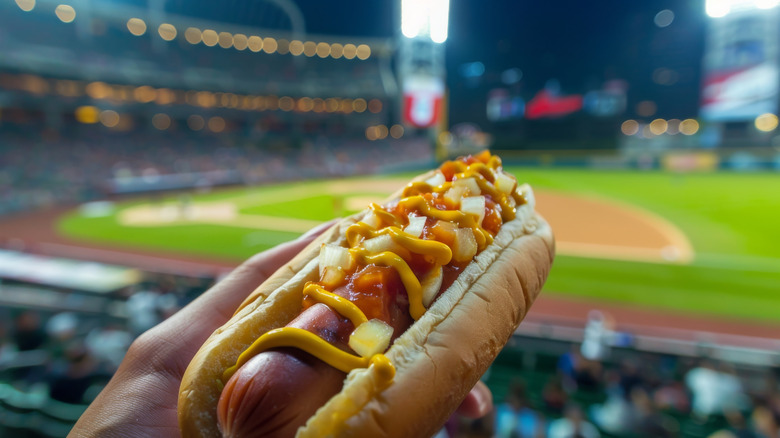 hand holding a hot dog in stadium stands overlooking baseball diamond