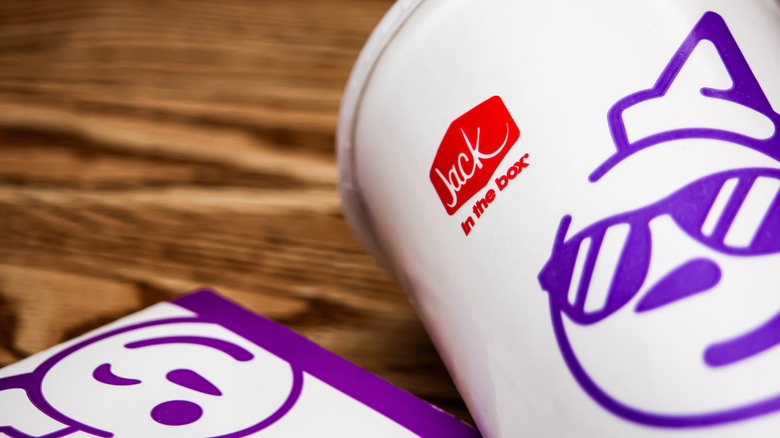 Closeup of a cup and food packaging with Jack In The Box logos, including the iconic clown logo wearing sunglasses and smirking