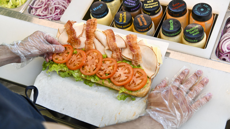 Subway sandwich on assembly line, open, person places tomato on sandwich