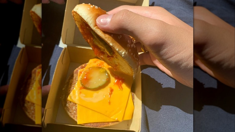 A hand lifting the top bun off a burger that contains only cheese, sauce, and pickles, no meat
