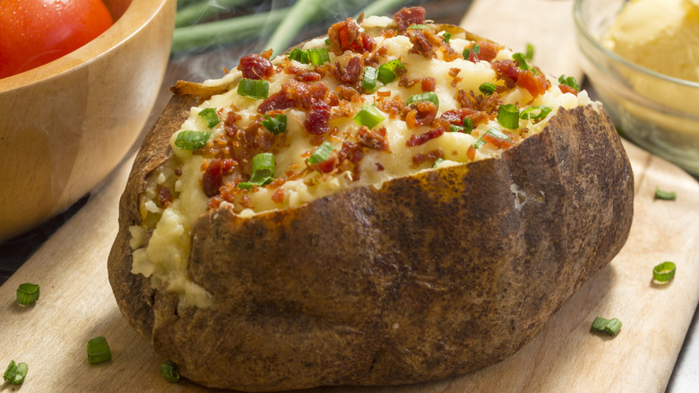 A large baked potato with onion and chives on a serving tray with bowls surrounding it
