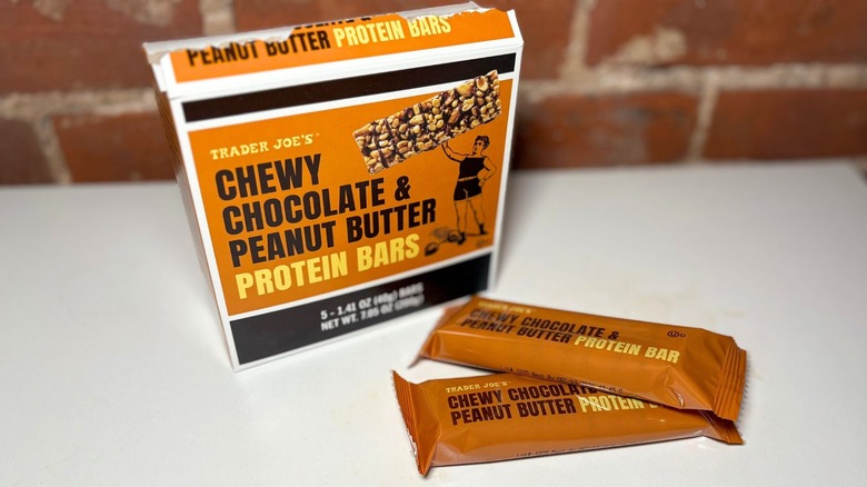A box of Chewy Chocolate & Peanut Butter Protein Bars next to two bars on a white countertop against a brick background