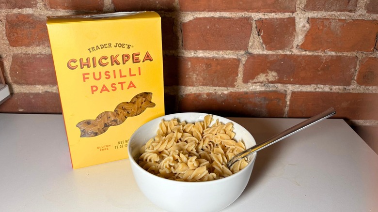 A box and white bowl of Trader Joe's Chickpea Fusilli Pasta on a white countertop against a brick background