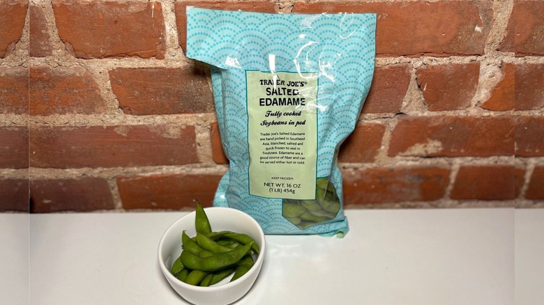 A package and white bowl of Lightly Salted Edamame on a white countertop against a brick background