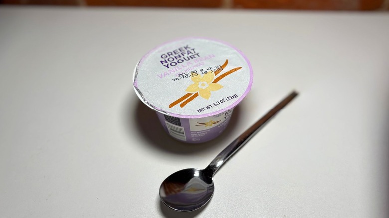 A package of Nonfat Vanilla Greek Yogurt on a white table against a brick background with a spoon